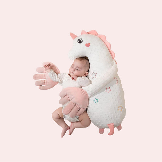 White unicorn-shaped plush with pink mane, horn, feet and oversized palms positioned around a baby
