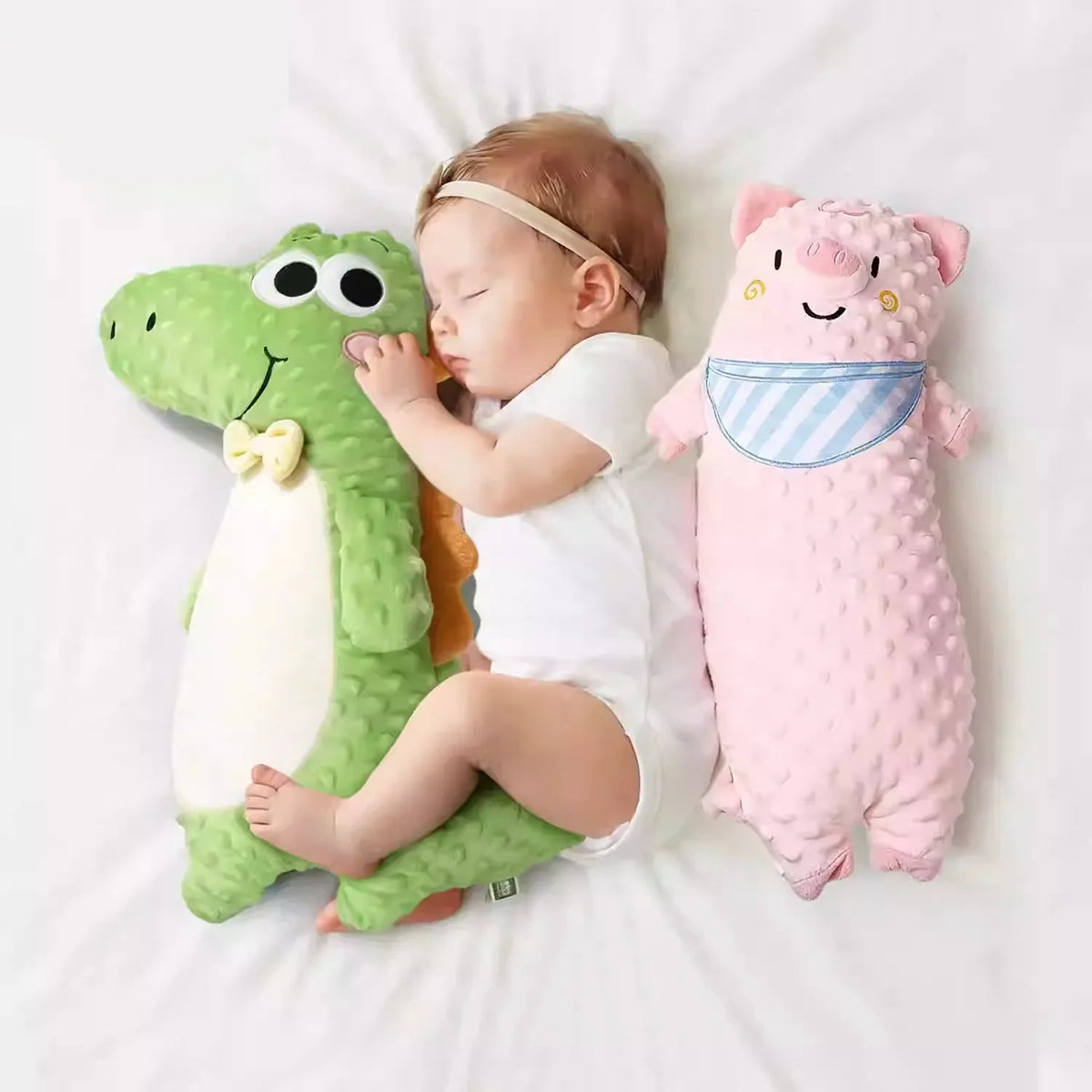 Sleeping newborn baby lying between a green minky dot crocodile plush and a pink pig soothing long pillow on white bedding.