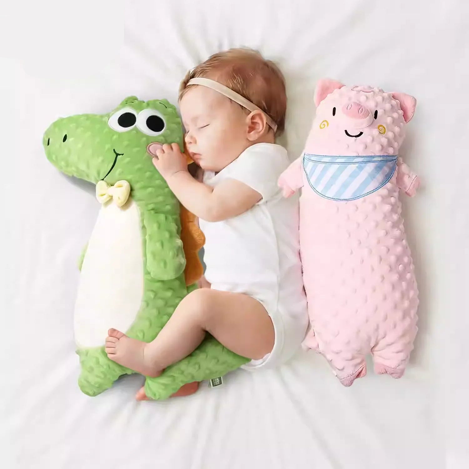 Sleeping newborn baby lying between a green minky dot crocodile plush and a pink pig soothing long pillow on white bedding.
